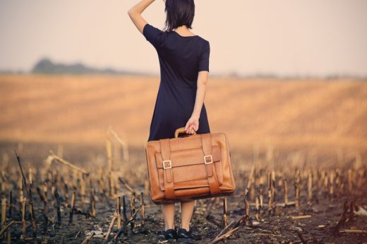 Captions for travel photo of a woman holding a suitcase in an open field alone.